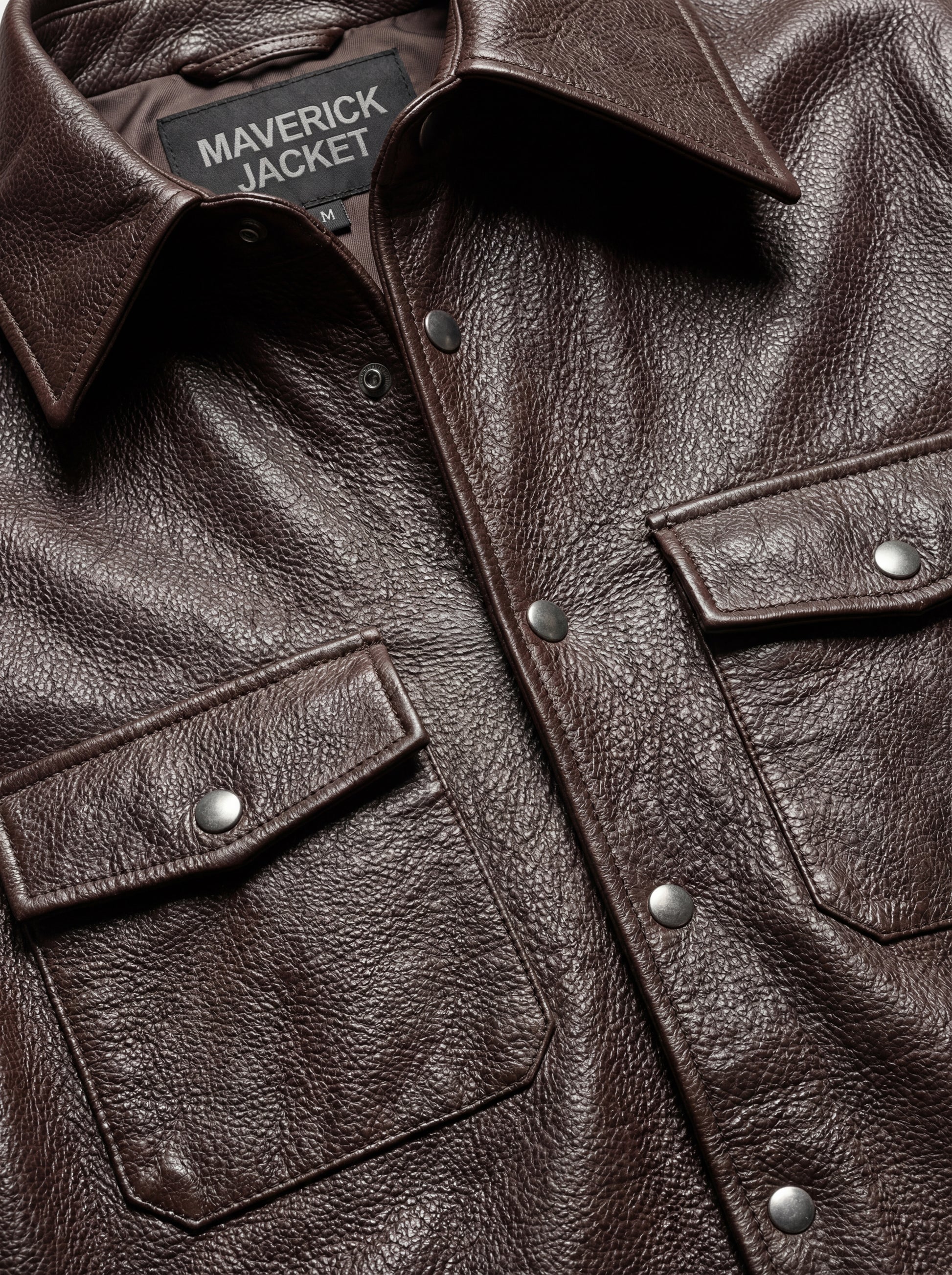 Macro shot of grained dark brown leather texture and silver snap button detail on chest pocket. The Garrison by Maverick Jacket.