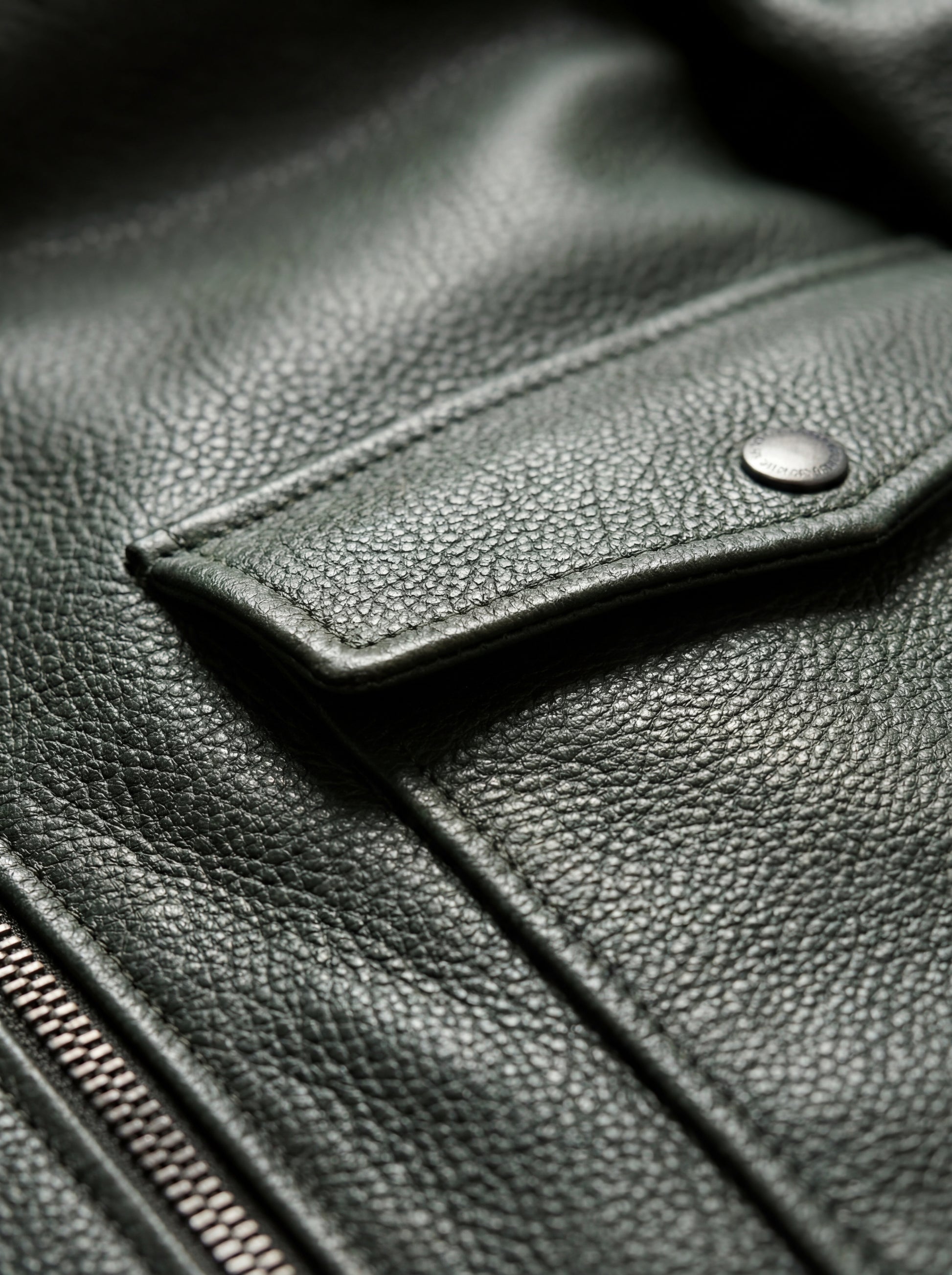 Macro shot of heavy pebbled green leather texture and metal snap button on pocket flap on bomber jacket. The Verdant by Maverick Jacket.