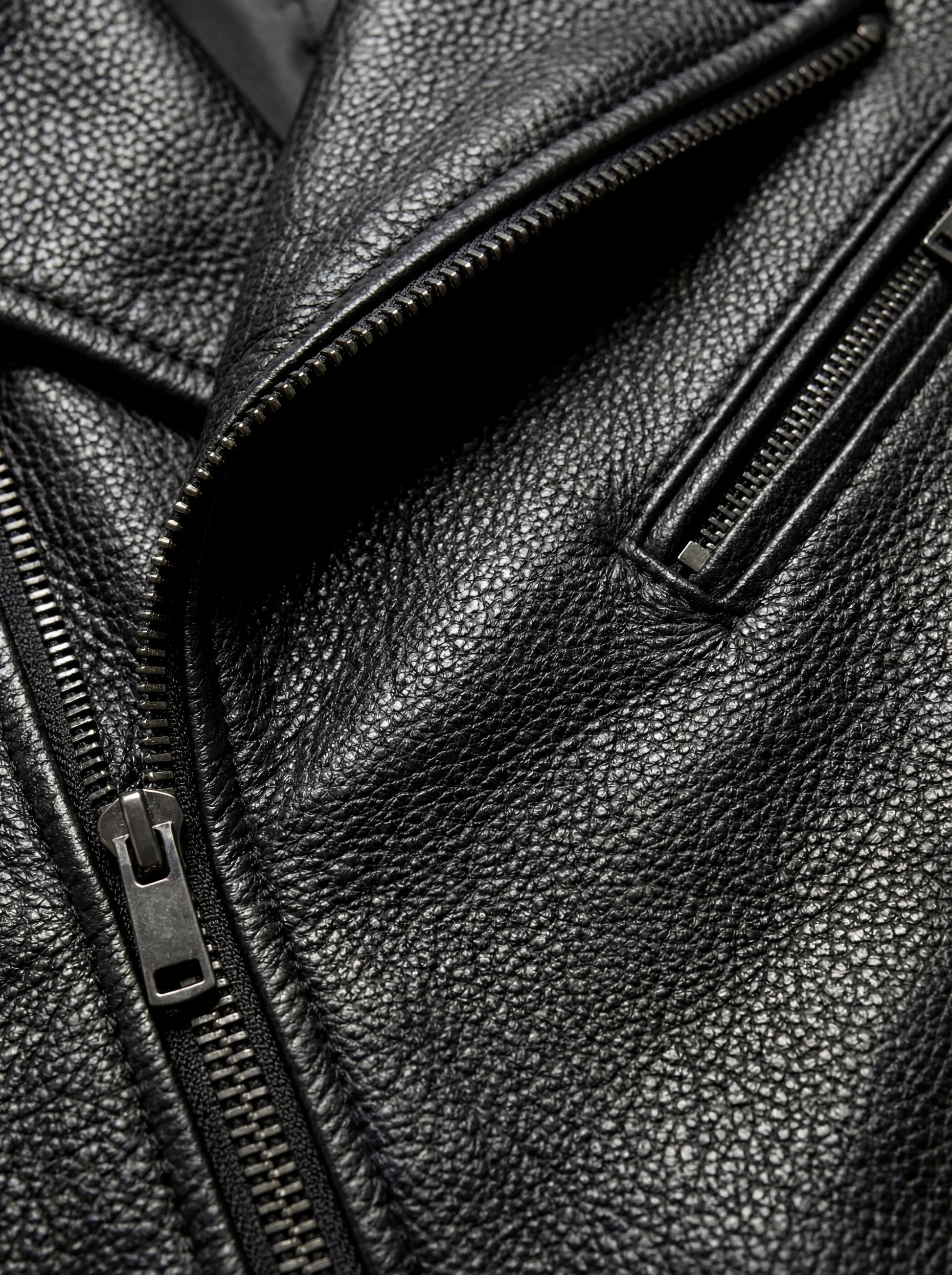 Macro shot of black pebbled leather grain texture and silver diagonal zipper pocket on biker jacket. The Night Rider by Maverick Jacket.
