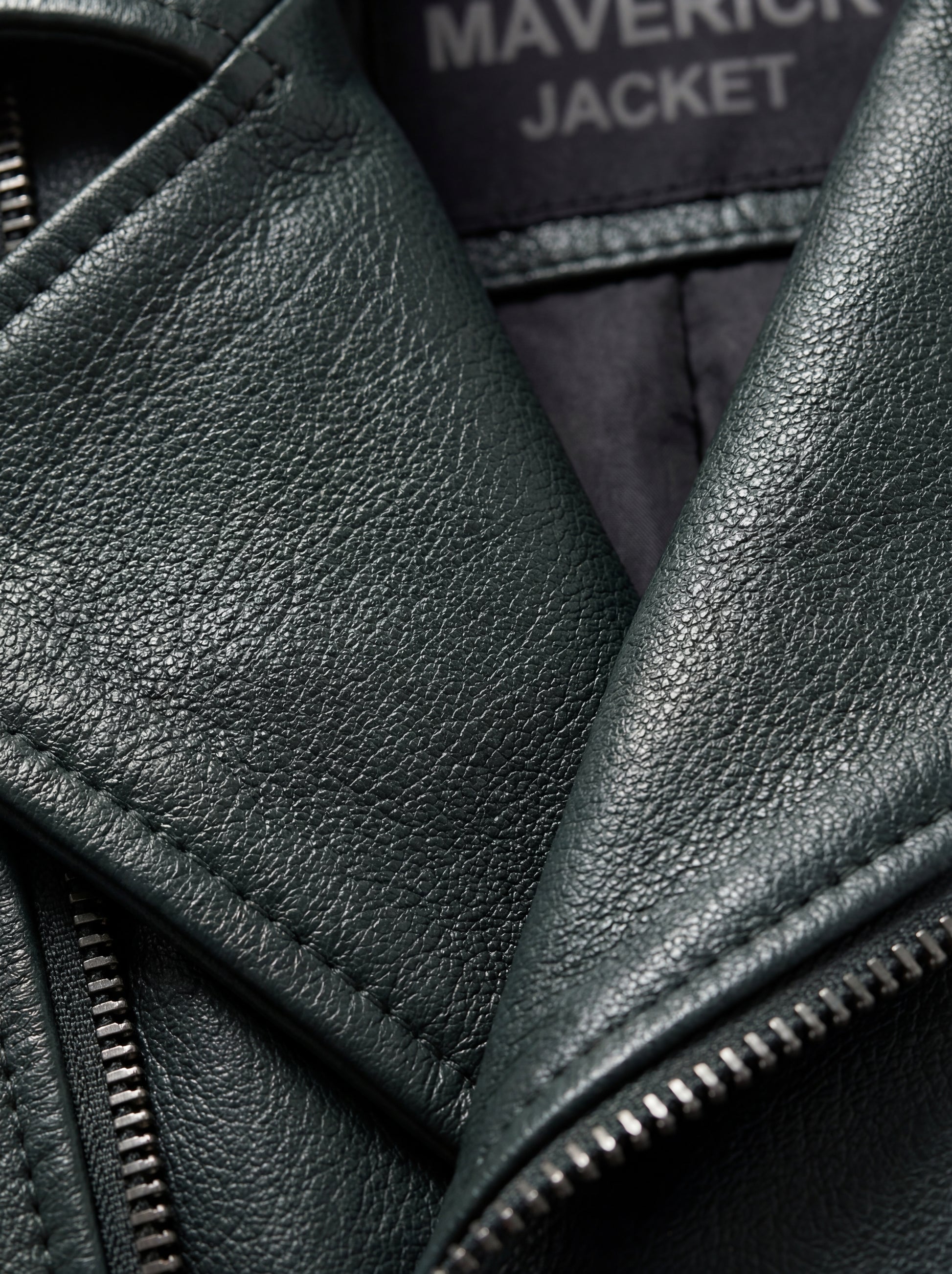 Macro shot of dark green pebbled leather grain texture and silver zipper hardware detail on biker jacket. The Rogue by Maverick Jacket.