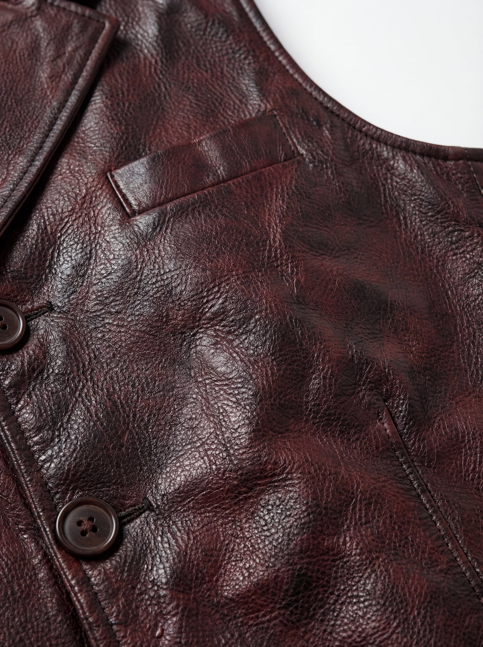 Macro shot of smooth burgundy leather texture and metal snap button hardware on vest. The Rook by Maverick Jacket.
