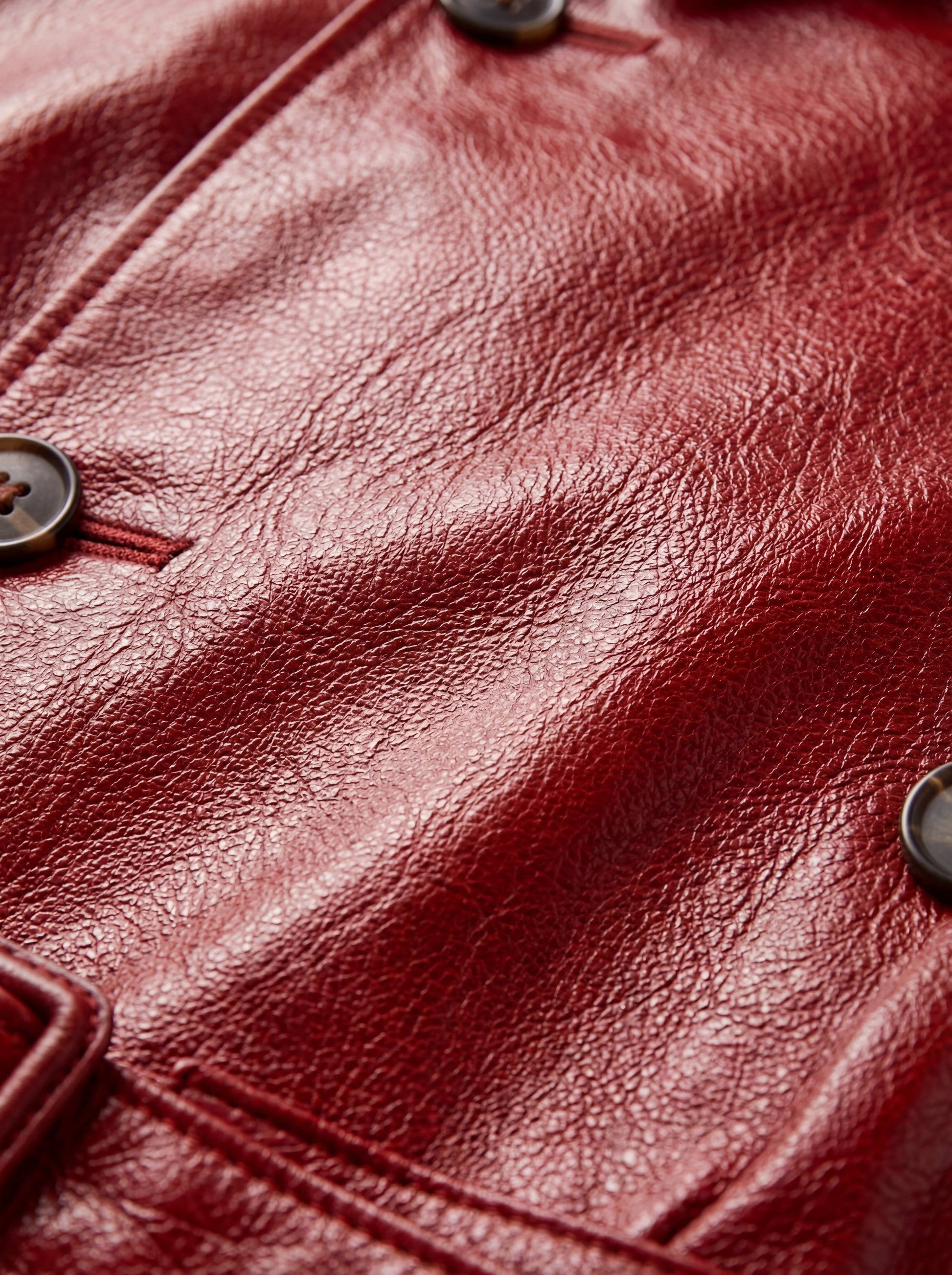 Macro shot of pebbled red leather texture and dark button detailing on trench coat. The Valerian by Maverick Jacket.