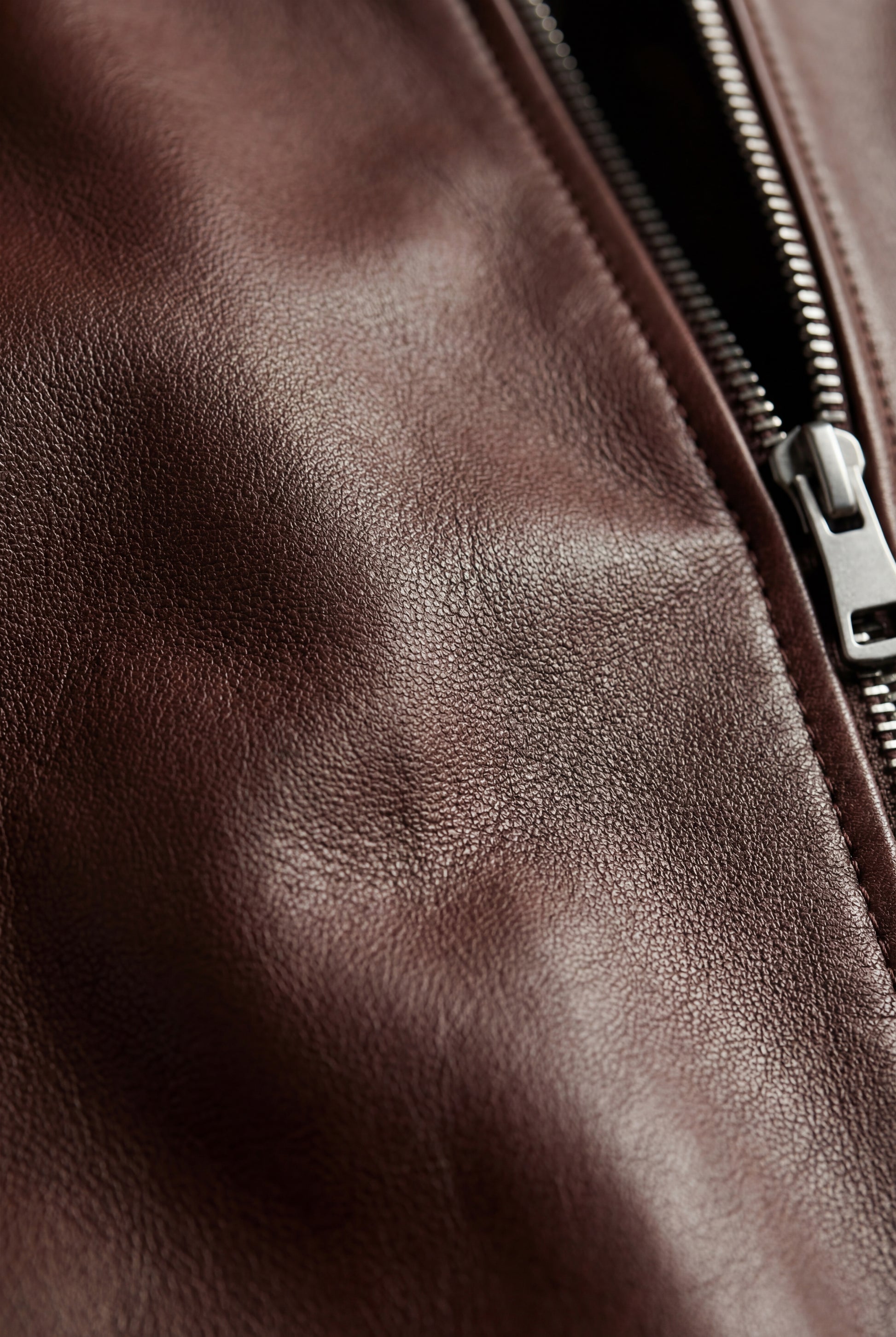 Macro shot of dark brown pebbled leather texture and silver zipper hardware on bomber jacket. The Ferran by Maverick Jacket.