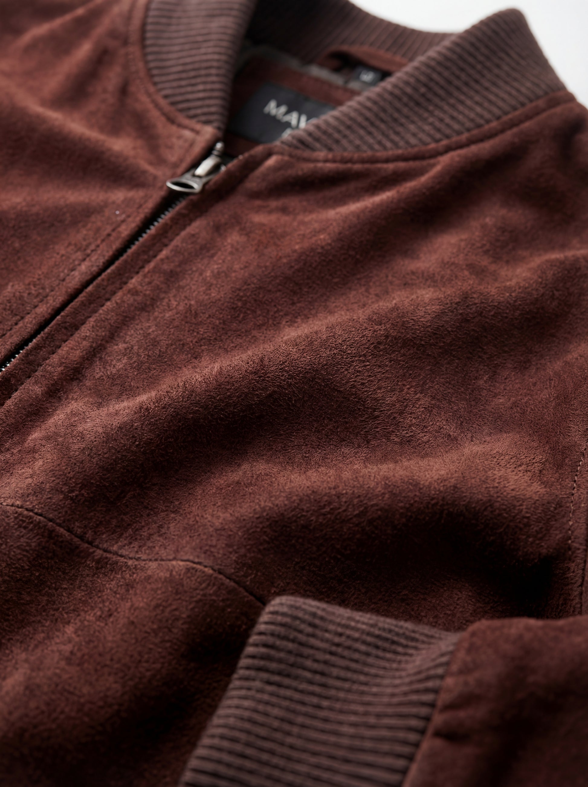 Macro shot of soft brown suede texture and metal zipper hardware on bomber jacket. The Miles by Maverick Jacket.