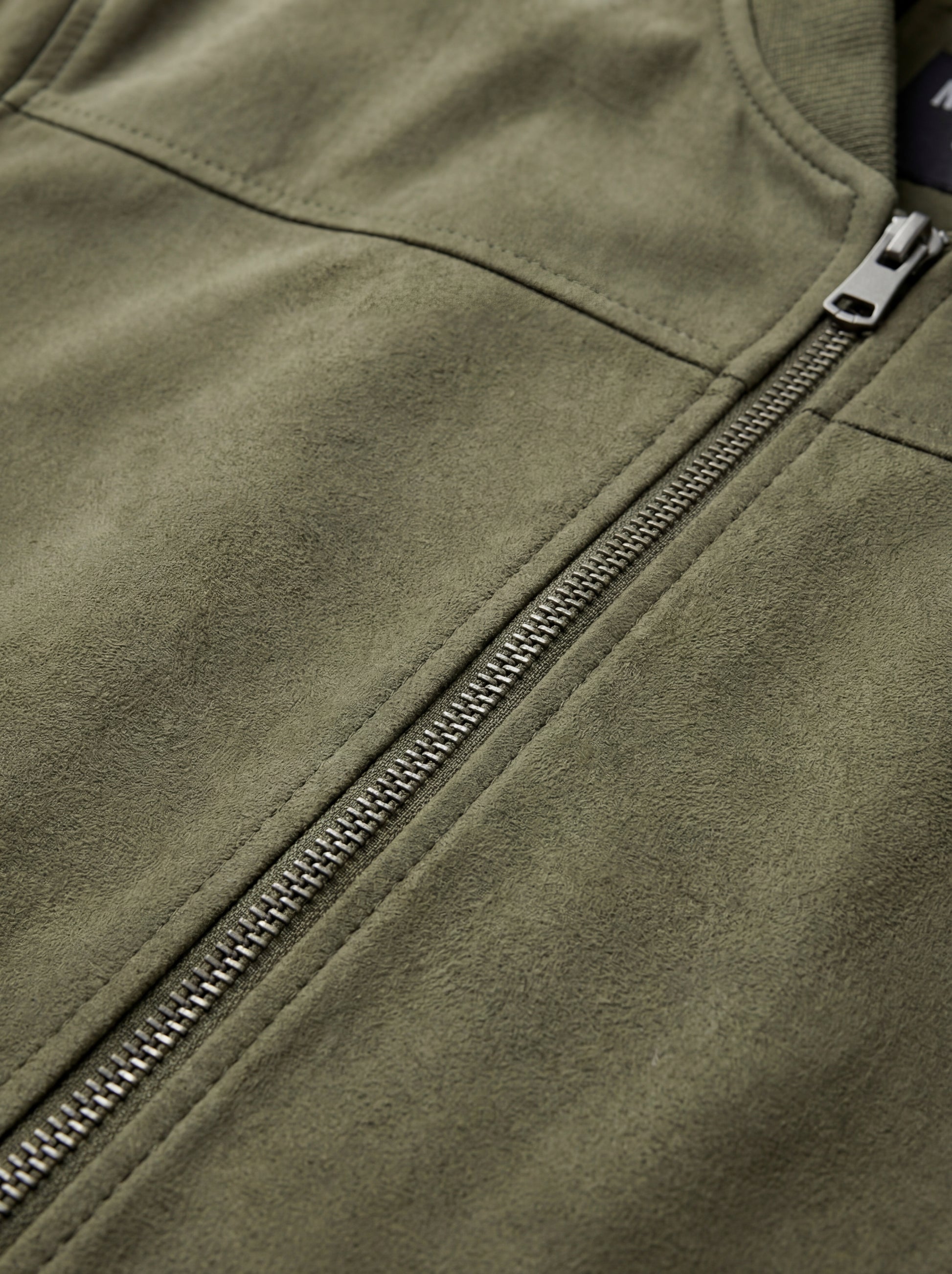 Macro shot of soft olive green suede texture and metal zipper hardware on bomber jacket. The Evan by Maverick Jacket.