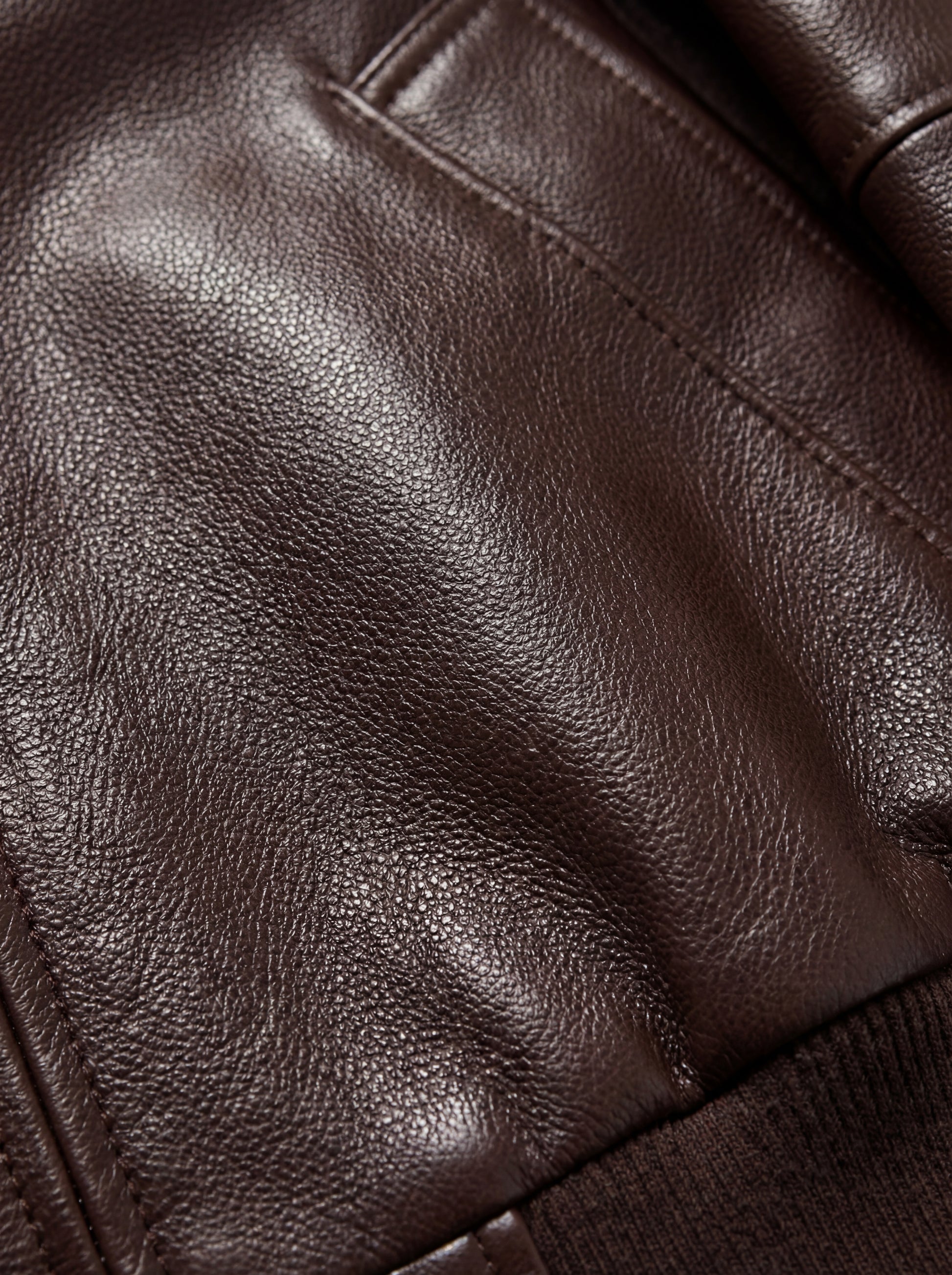Macro shot of grained brown leather texture and tonal stitching detail on bomber jacket. The Isla by Maverick Jacket.