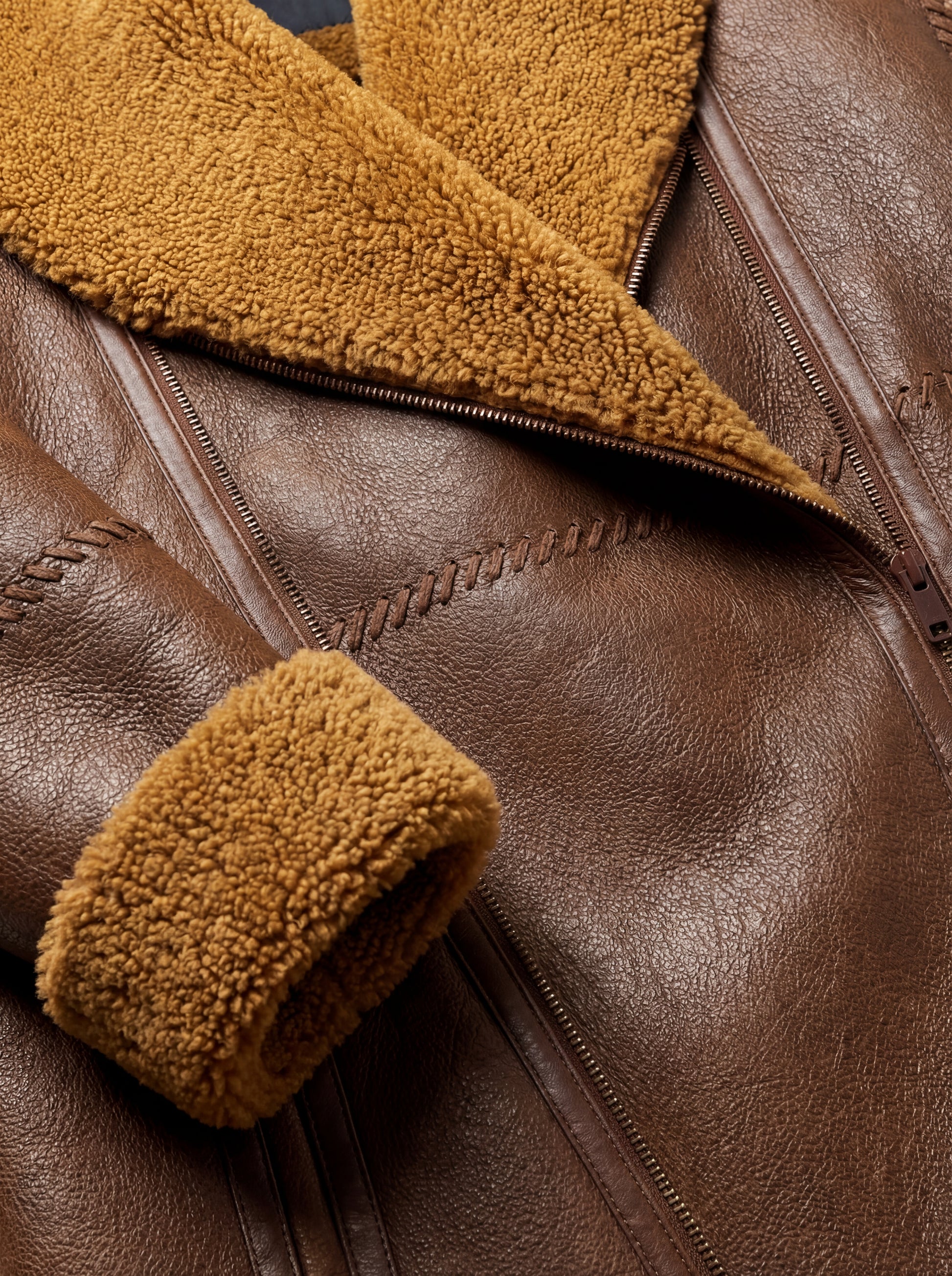 Macro shot of pebbled brown leather texture and decorative whipstitch detailing on shearling jacket. Maverick Greta.