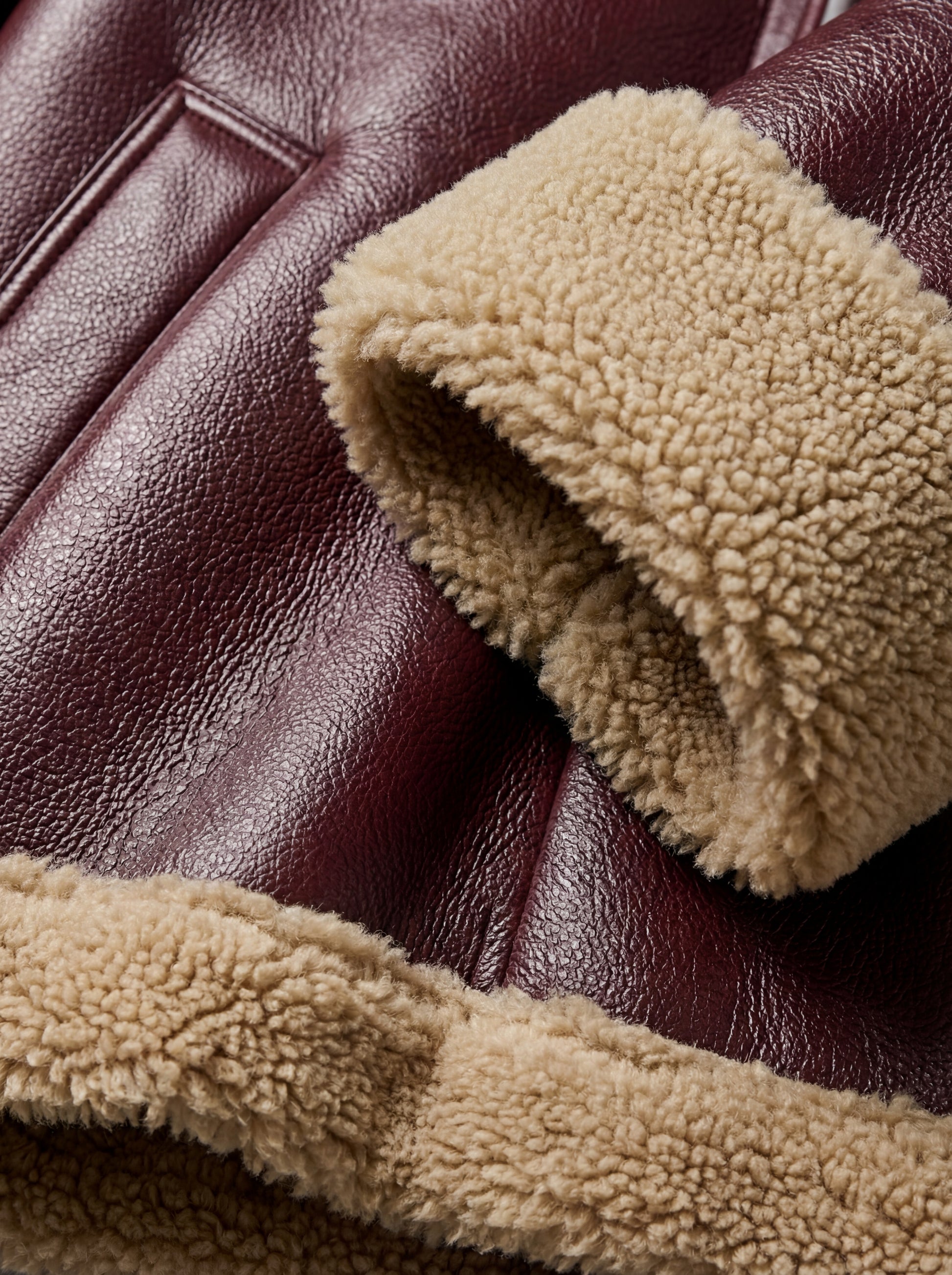 Macro shot of pebbled burgundy leather texture and thick beige shearling cuff on jacket. Maverick Faye.