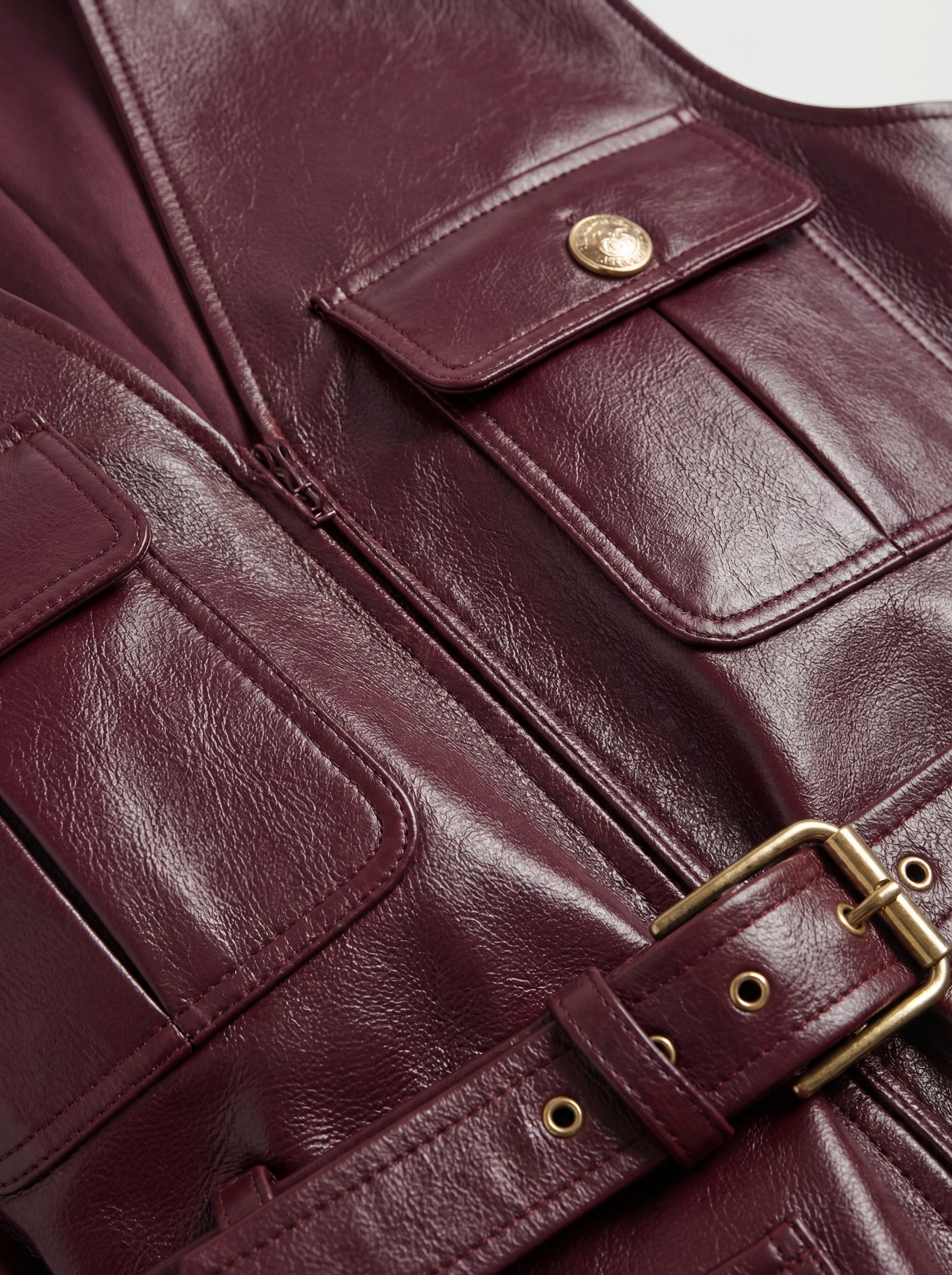 Macro shot of burgundy leather grain texture and gold embossed button detail on utility vest. The Victoria by Maverick Jacket.