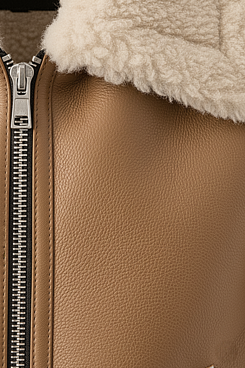 Macro shot of pebbled tan leather texture and silver metal zipper hardware on shearling vest. The Clara by Maverick Jacket.