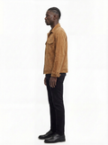 A man in profile wearing a tan suede jacket, black trousers, and black shoes, standing against a plain white studio background.